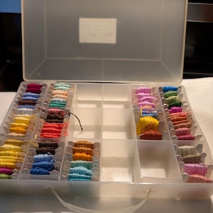 May include: A translucent plastic storage box with a hinged lid containing numerous embroidery floss skeins wound onto small cardboard bobbins. The bobbins are organized in divided compartments and feature a wide variety of colors including blue, yellow, pink, green, brown, and white. The box has a handle on the front.