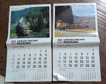 May include: Two vintage Union Pacific Railroad calendars. The July 1994 calendar features a train on a track through a forest. The July 1995 calendar shows a train near a mountain. Both calendars have a July calendar grid.
