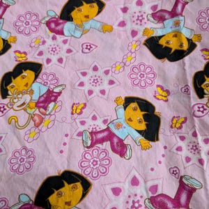 May include: Pink fabric featuring a repeating pattern of a cartoon character with black hair, wearing a light blue shirt and pink pants. The design includes flowers, stars, and butterflies. The character is in various poses.