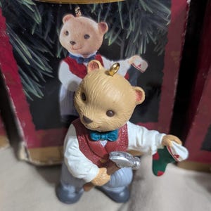 May include: A Christmas ornament featuring a tan teddy bear wearing a red vest, white shirt, and blue bow tie. The bear holds a silver hammer and a green and red stocking. A second bear is visible in the background.