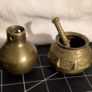 May include: Two antique brass items: a small, round bottle with a hole and a small brass mortar and pestle. The mortar has a decorative pattern around the base. Both items have a tarnished, aged appearance.