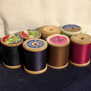 May include: A collection of vintage wooden spools of thread in various colors, including black, navy blue, brown, and burgundy. The spools have colorful paper labels with text and are arranged on a dark blue surface.