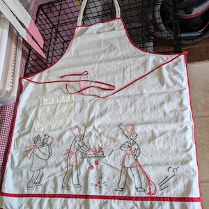 May include: A white kitchen apron featuring red trim along the edges and a single pocket. The apron is decorated with black and red embroidered line art illustrations of three figures performing household tasks, including cooking and sweeping.