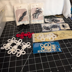May include: A collection of various frog closures and decorative buttons in black, red, and white. Some are packaged, while others are loose. The closures feature intricate designs, including floral and looped patterns. The image includes the text "FROG CLOSURE".