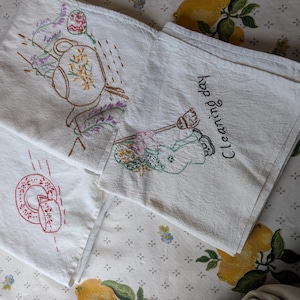 May include: Three white fabric towels featuring embroidered designs. One towel shows a teapot with lavender, another displays a red teacup and saucer, and the third depicts a figure holding a broom with the text Cleaning day embroidered.