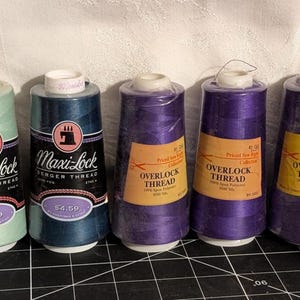 May include: Five spools of sewing thread in various colors. Two spools are labeled Maxi-Lock Serger Thread, and three are labeled Overlock Thread. The spools are in shades of teal, navy, and purple.