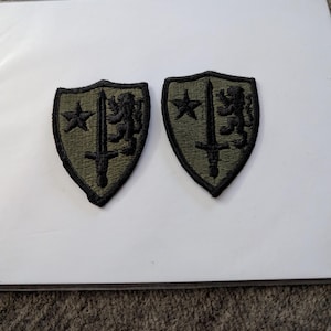 May include: Two shield-shaped embroidered patches in olive green with a black border. Each patch features a black embroidered design consisting of a five-pointed star on the left, a vertical sword in the center, and a rampant lion on the right side.
