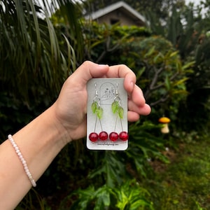May include: Cherry earrings with red, glossy beads and green leaf accents. The earrings are held on a small, rectangular card with the text "lunarcraftydesign.com". The earrings are suspended from silver-colored hooks.
