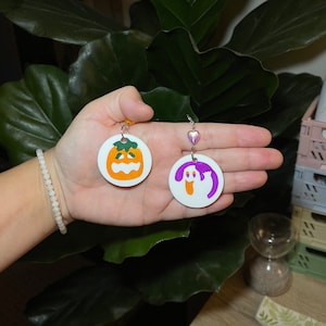 K&ouml;nnte beinhalten: Ein Paar Halloween-Ohrringe. Ein Ohrring zeigt einen wei&szlig;en Kreis mit einem lila Geist, der andere Ohrring zeigt einen wei&szlig;en Kreis mit einem orangefarbenen K&uuml;rbis.