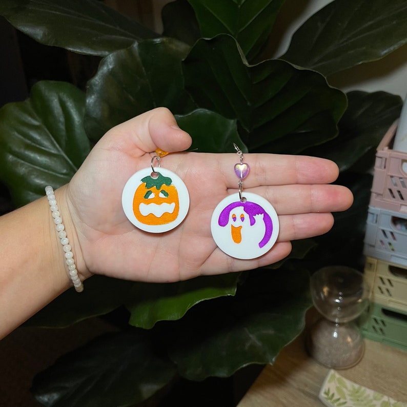 K&ouml;nnte beinhalten: Ein Paar Halloween-Ohrringe. Ein Ohrring zeigt einen wei&szlig;en Kreis mit einem lila Geist, der andere Ohrring zeigt einen wei&szlig;en Kreis mit einem orangefarbenen K&uuml;rbis.