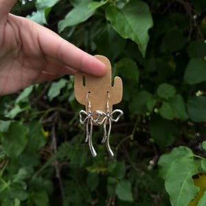 Könnte beinhalten: Ein Paar silberfarbene Metallohrringe in Form von Schleifen mit langen, fließenden Bändern. Die Ohrringe sind an einer hellbraunen, kaktusförmigen Pappkarte befestigt und verfügen über einen einfachen Hakenverschluss für Ohrlöcher.