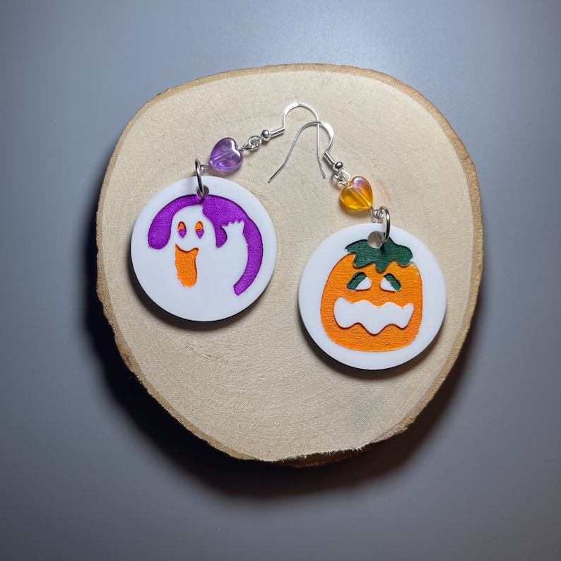 K&ouml;nnte beinhalten: Ein Paar Halloween-Ohrringe. Ein Ohrring zeigt einen wei&szlig;en Geist mit einer violetten Umrandung, der andere Ohrring zeigt einen wei&szlig;en K&uuml;rbis mit einem orangefarbenen Gesicht und einem gr&uuml;nen Stiel.