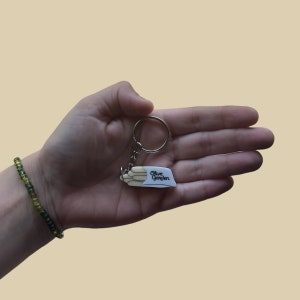 May include: A white keychain with a depiction of a breadstick bundle and the text "Olive Garden" on it. The keychain is attached to a silver key ring.