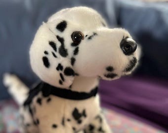 May include: A plush Dalmatian dog toy with white fur and black spots. The stuffed animal features dark brown plastic eyes, a black plastic nose, and a simple black fabric collar around its neck. The toy is depicted in a seated position.