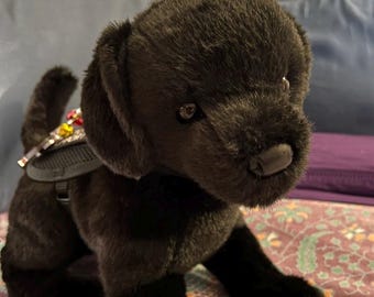 May include: Black plush toy dog wearing a black harness with colorful embellishments. The dog is sitting on a patterned purple and green fabric.