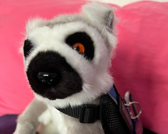 May include: A plush lemur toy with black and white markings, wearing a black harness with a mesh pouch and a silver key ring. The lemur has a white face with black markings around the eyes and a black nose. One eye is orange.