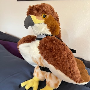 Puede incluir: Un halcón de peluche marrón y blanco con patas amarillas y garras negras. El halcón lleva un arnés negro con una hebilla plateada.
