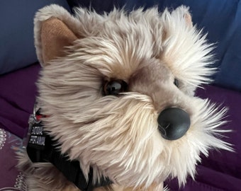 Peut inclure: Une peluche en forme de chien avec une fourrure hirsute beige et fauve, des yeux marron foncé et un nez noir. Elle porte un collier noir avec une étiquette rectangulaire portant l'inscription EMOTIONAL SUPPORT ANIMAL en lettres capitales blanches.