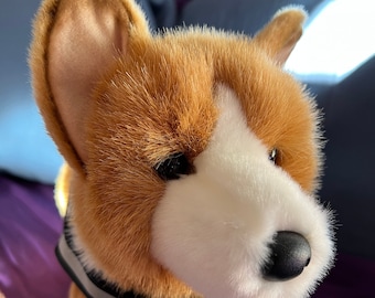 Peut inclure: Un jouet en peluche représentant un corgi brun et blanc avec un collier réfléchissant noir et gris. Le jouet est posé sur une surface violette.