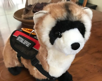 Pode incluir: Um furão de pelúcia vestindo um colete de animal de serviço preto e vermelho com o texto "Emotional Support". O furão é marrom, branco e preto com olhos pretos.