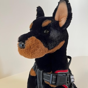 May include: A plush Doberman Pinscher dog with black fur, brown markings, and a black and gray harness. The harness has a red accent and the words "EMOTIONAL SUPPORT" printed on it. The dog has a soft, plush appearance.