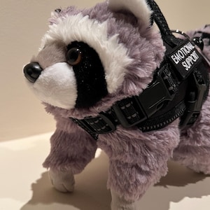 Puede incluir: Un peluche de mapache con pelaje gris y blanco, que lleva un arnés negro de estilo táctico. El arnés cuenta con un parche negro con el texto blanco EMOTIONAL SUPPORT, correas ajustables con hebillas de plástico y un asa en la parte superior.
