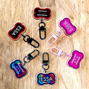 Puede incluir: Una colección de llaveros coloridos en forma de hueso. Los llaveros presentan la palabra "ESSA" en varios colores, junto con el texto "EMOTIONAL SUPPORT STUFFED ANIMAL". Cada llavero tiene un cierre metálico.