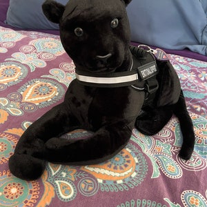 May include: Black plush panther toy wearing a black and white harness with the text "Emotional Support" on it. The toy is lying on a purple and orange patterned bedspread.
