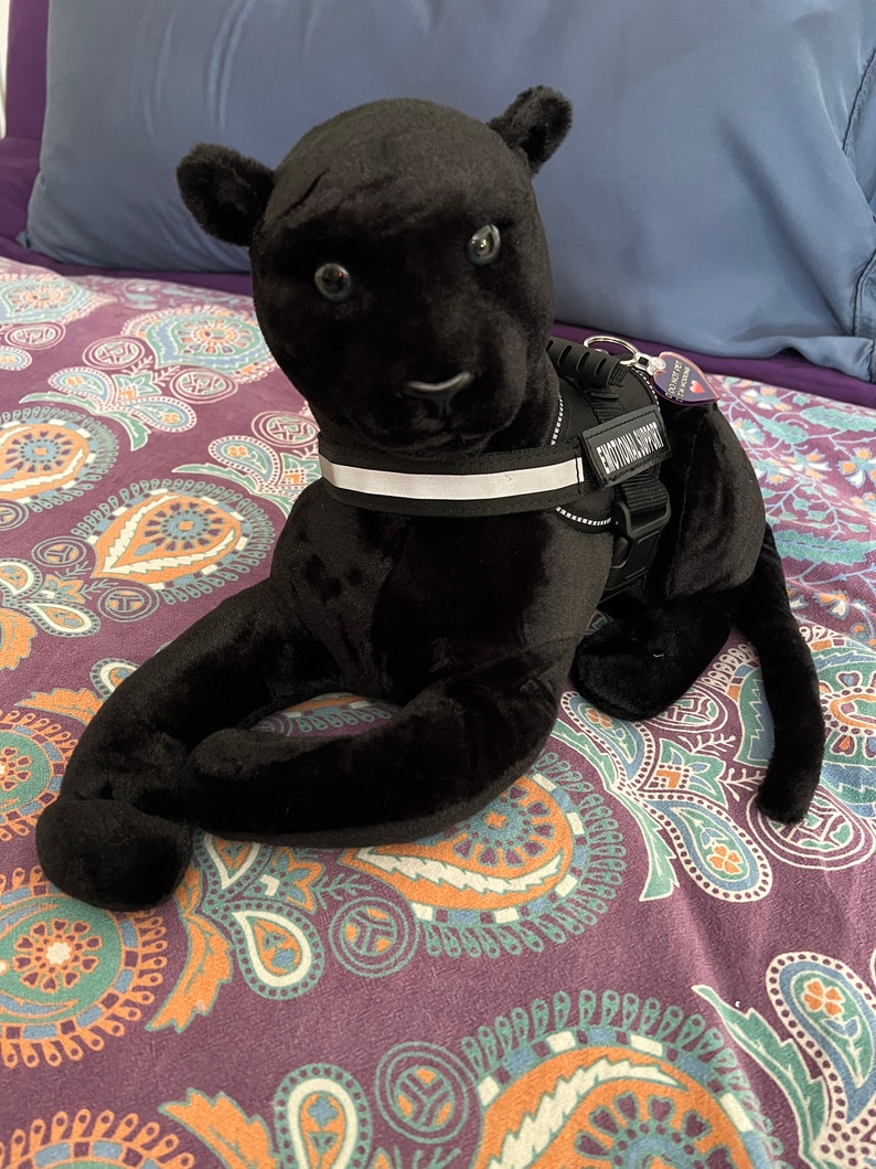 May include: Black plush panther toy wearing a black and white harness with the text "Emotional Support" on it. The toy is lying on a purple and orange patterned bedspread.