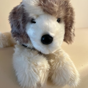 Könnte beinhalten: Ein Plüschhund mit weichem, langflorigem Fell in Creme- und Hellbrauntönen. Das Stofftier hat ein weißes Gesicht, braune Ohren und eine schwarze Kunststoffnase. Es ist in einer liegenden Position mit nach vorne ausgestreckten Pfoten dargestellt.
