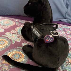 May include: Black plush toy dog wearing a black harness with the text "Emotional Support" and a red heart-shaped tag that says "Do Not Pet I'm Working".