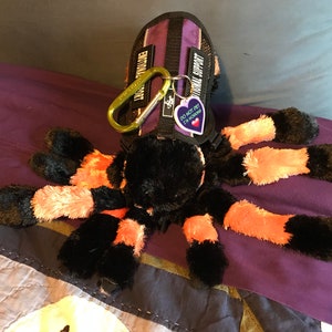 May include: A black and orange plush spider toy with a purple and black harness and a tag that reads "Do Not Pet I'm a Spooder".