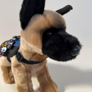 May include: A plush German Shepherd dog toy featuring tan fur with a black muzzle and ears. The toy wears a blue and black harness with a small metal tag shaped like a paw print and a yellow gem charm attached to the harness strap.
