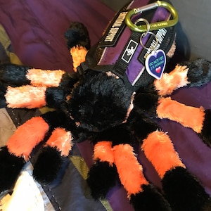 May include: A black and orange plush spider toy wearing a purple and black harness with the text "Emotional Support" and a tag that says "Do Not Pet I'm Working".