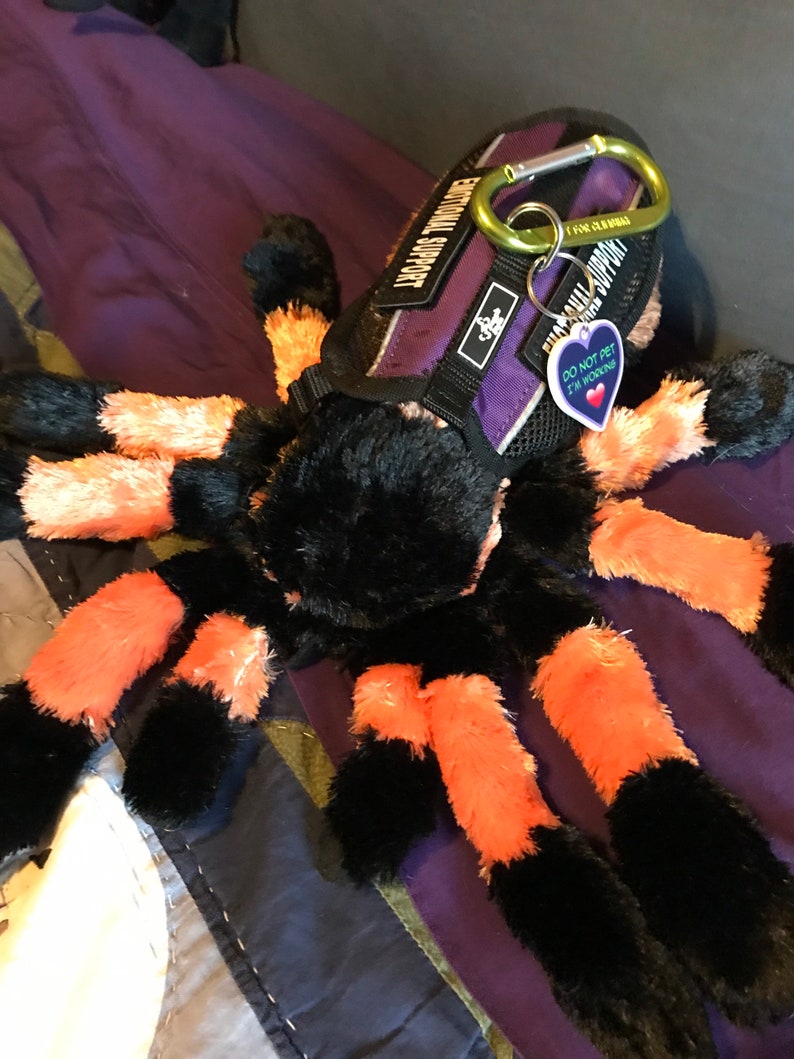 May include: A black and orange plush spider toy wearing a purple and black harness with the text "Emotional Support" and a tag that says "Do Not Pet I'm Working".
