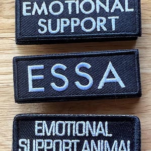Może przedstawiać: Trzy prostokątne czarne naszywki z białym haftowanym tekstem. G&oacute;rna naszywka ma napis EMOTIONAL SUPPORT, środkowa ESSA, a dolna EMOTIONAL SUPPORT ANIMAL. Każda naszywka posiada czarną obw&oacute;dkę oraz widoczną fakturę materiału.