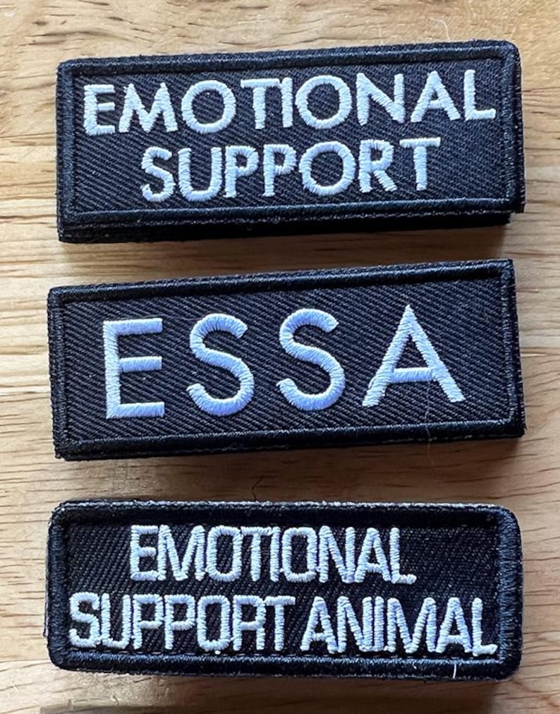 Może przedstawiać: Trzy prostokątne czarne naszywki z białym haftowanym tekstem. G&oacute;rna naszywka ma napis EMOTIONAL SUPPORT, środkowa ESSA, a dolna EMOTIONAL SUPPORT ANIMAL. Każda naszywka posiada czarną obw&oacute;dkę oraz widoczną fakturę materiału.