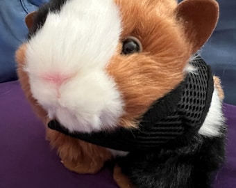 May include: A plush guinea pig toy featuring a tri-color coat of white, brown, and black fur. The stuffed animal has a soft white face with a pink nose, dark eyes, and rounded ears. It is wearing a black mesh harness around its midsection.