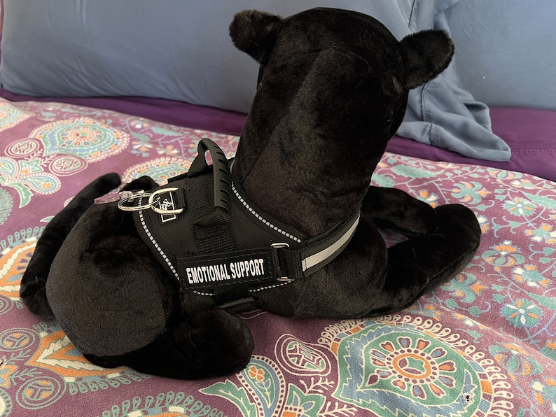 May include: Black plush dog wearing a black harness with the words "Emotional Support" printed on it. The dog is lying on a purple and green patterned bedspread.