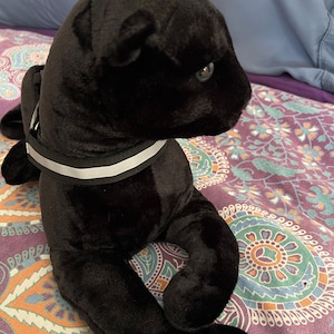 May include: A black plush panther toy with a white reflective collar. The panther is lying on a patterned fabric.