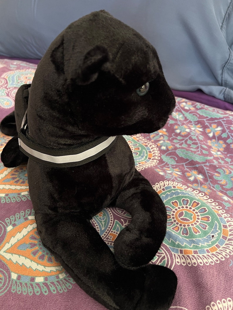 May include: A black plush panther toy with a white reflective collar. The panther is lying on a patterned fabric.