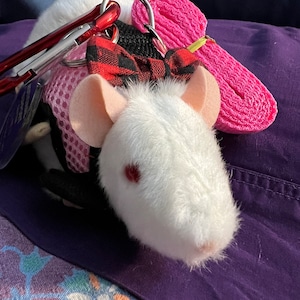 May include: A white plush rat toy wearing a black and pink mesh harness with a red and black buffalo plaid bow. The harness features a metal ring attached to a red carabiner and a long, folded pink nylon leash strap for secure handling and control.