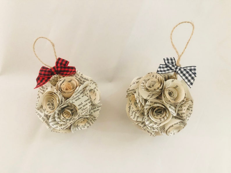 May include: Two handmade paper ornaments, each crafted from book pages and shaped into a sphere. Each ornament features paper roses and a small gingham bow. One bow is red and black, the other is black and white.
