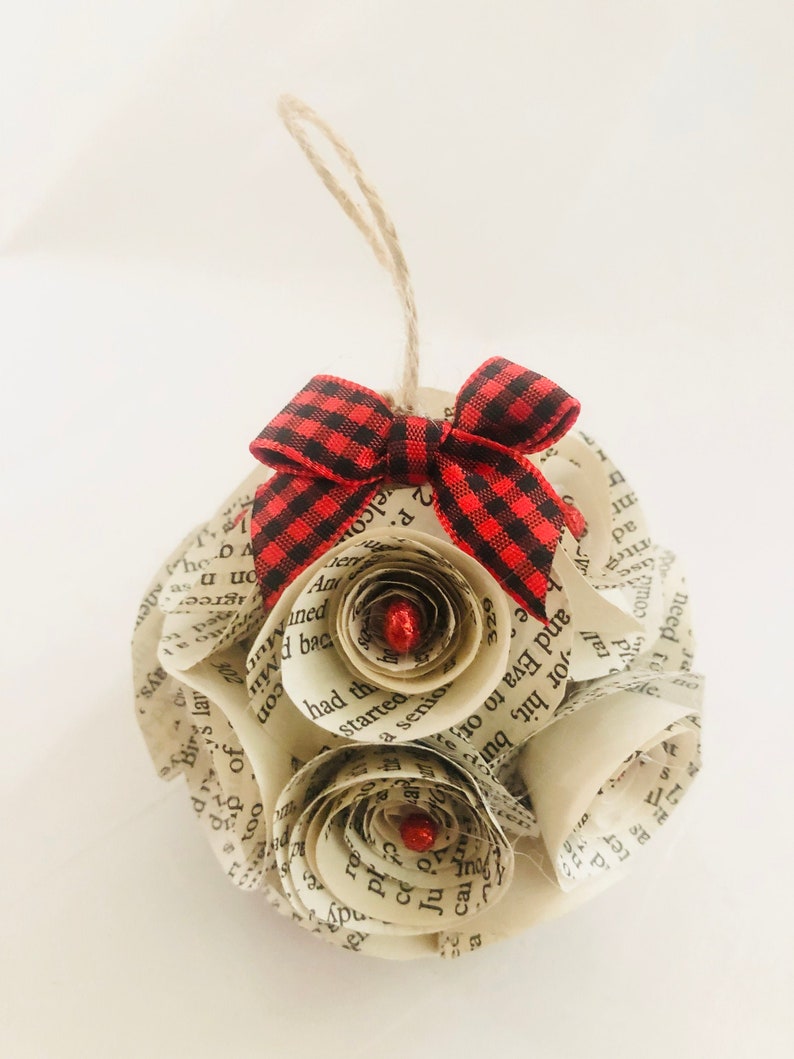 May include: A handmade ornament crafted from book pages, shaped into a sphere with paper roses. It features a red and black plaid bow and a twine loop for hanging. The roses have red glitter centers.