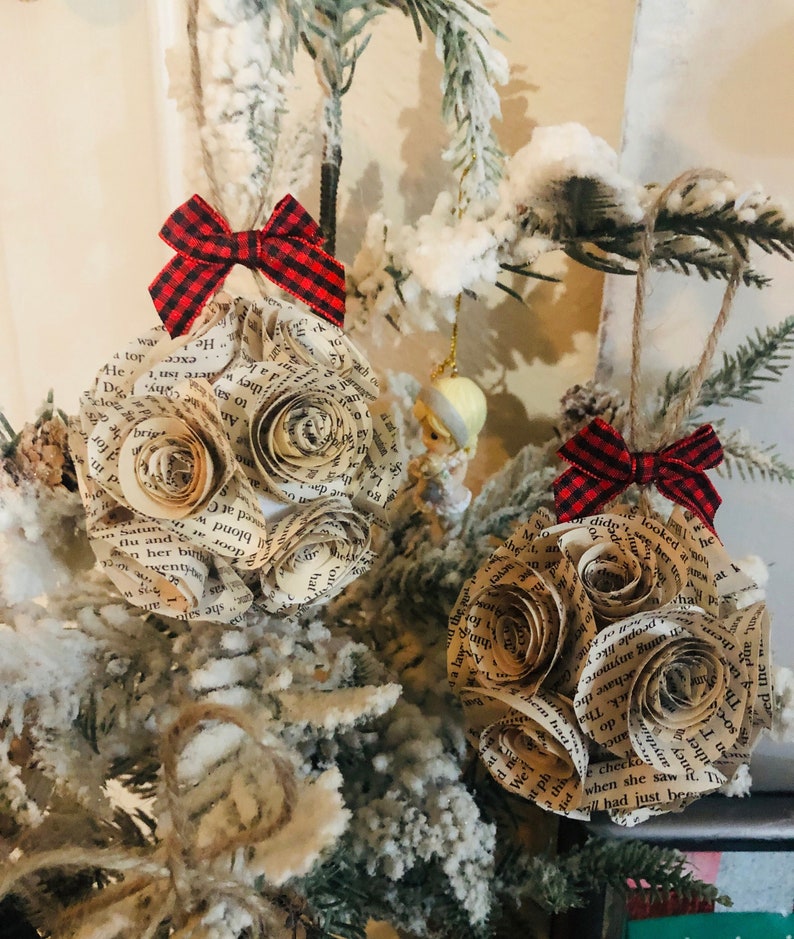 May include: Two handmade Christmas ornaments. Each ornament is a sphere made of rolled book pages, resembling roses. They are topped with a red and black plaid bow and suspended by twine. The ornaments are displayed on a snowy evergreen branch.