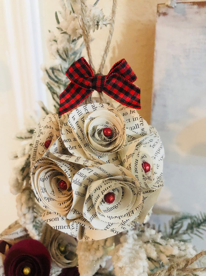 May include: A handmade Christmas ornament featuring paper roses crafted from book pages. The ornament has red glitter accents and a red and black plaid bow. It hangs from a twine loop, perfect for holiday decor.