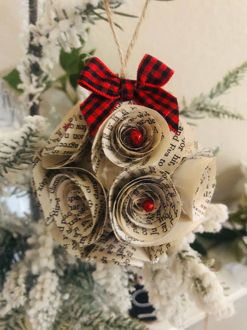 May include: A handmade Christmas ornament made from book pages, shaped into a sphere with paper roses. It has a red and black plaid bow and a string for hanging. The roses have red glitter centers.