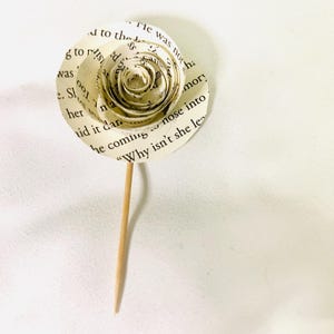 May include: A handmade paper rose crafted from book pages, featuring visible text. The flower has a spiral center and is attached to a wooden skewer. The petals display printed text, creating a unique, literary-themed decorative item.