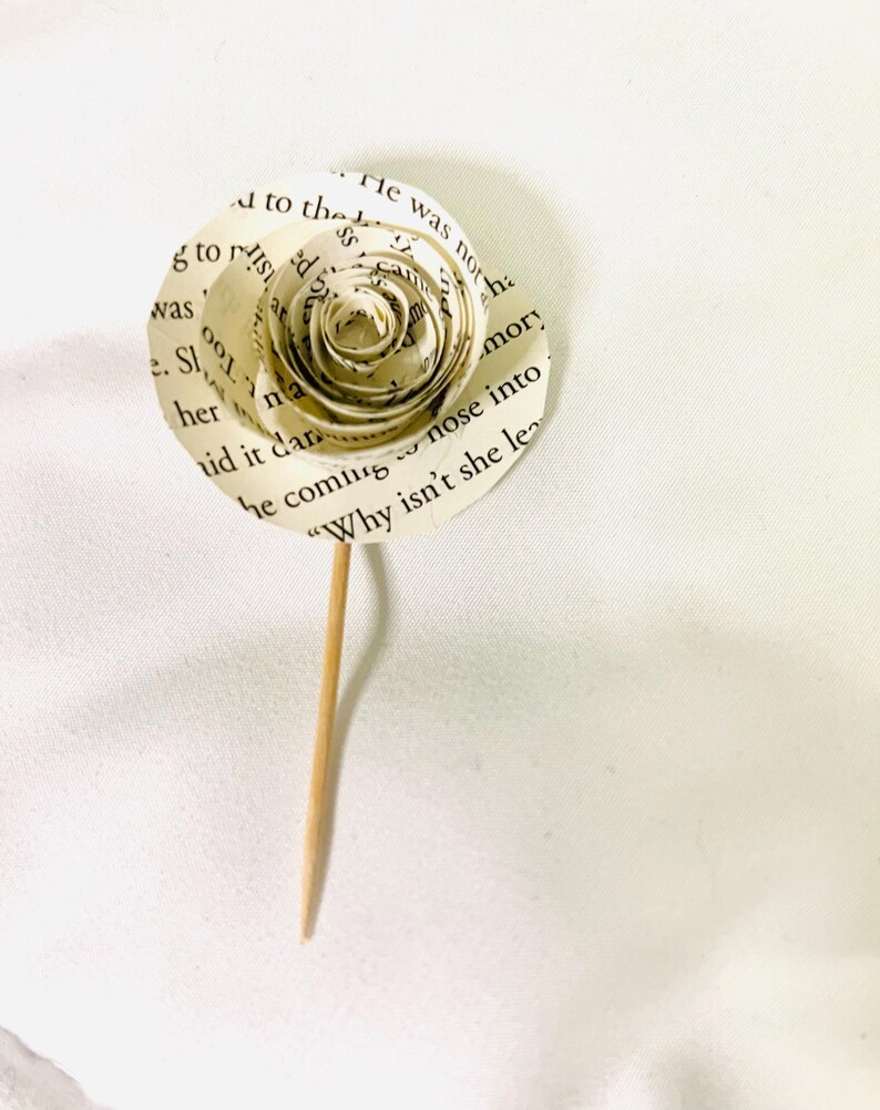 May include: A handmade paper rose crafted from book pages, featuring visible text. The flower has a spiral center and is attached to a wooden skewer. The petals display printed text, creating a unique, literary-themed decorative item.