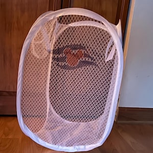 May include: White mesh pop-up laundry hamper with a white trim and handles. The hamper has a cartoon Mickey Mouse design in the center. The hamper is open and sitting on a wooden floor.
